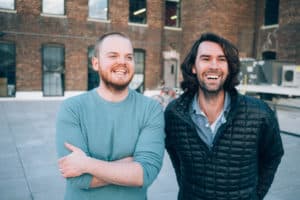 Zack Akers, a white man with short hair and a short beard wearing a gray-blue sweater crosses his arms and smiles. Skip Bronkie, a white man with longer brown hair and a short beard dressed in a blue button-down shirt and black jacket, has his arms behind him and smiles. The two stand in front of a brick building.