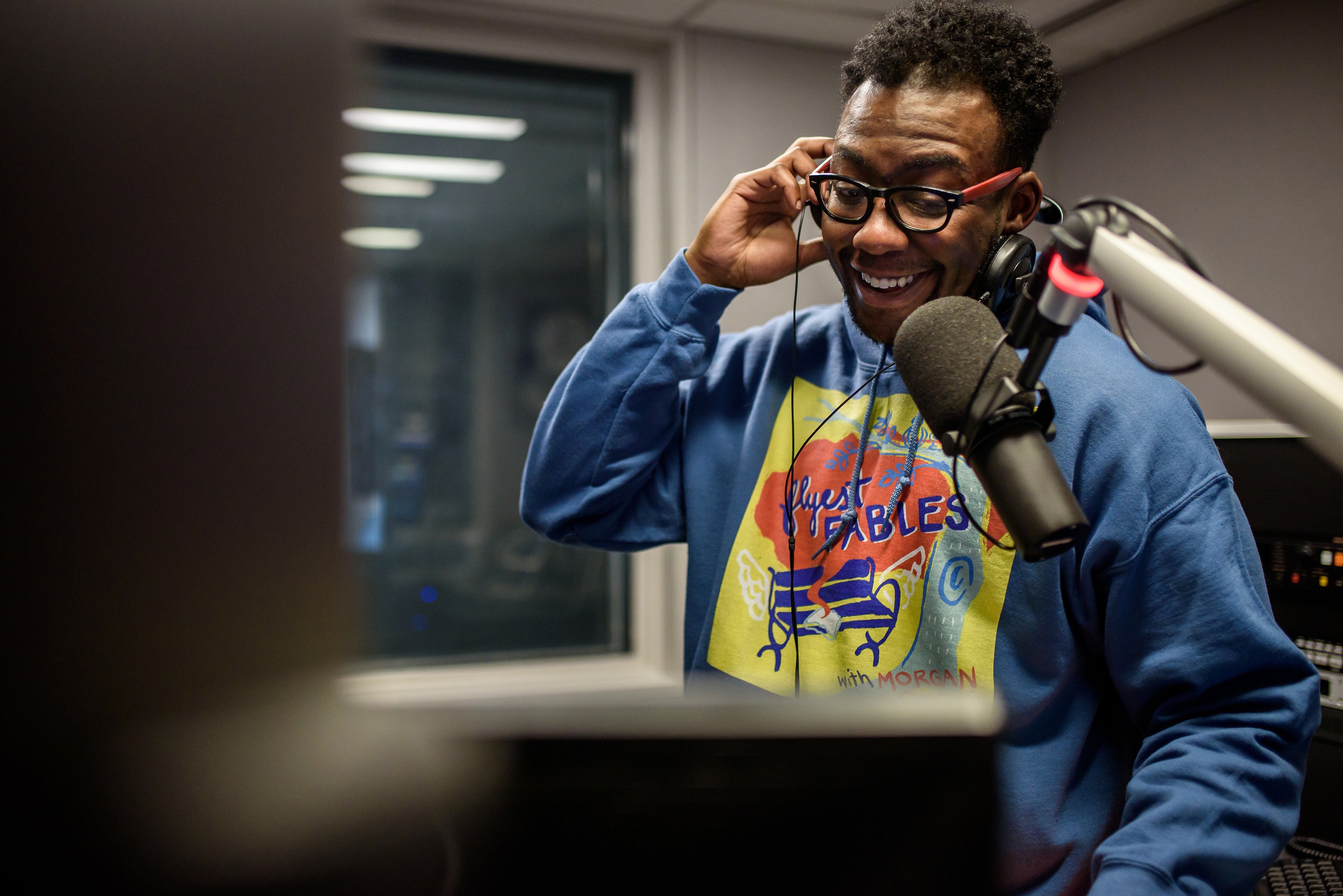 Morgan Givens in front of a microphone in a sound booth, his hand holding his headphones to his ears. He is wearing glasses and a blue sweatshirt with the logo for his podcast, Flyest Fables